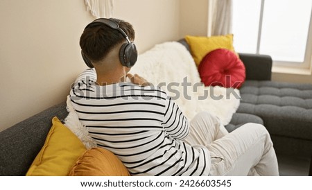 Attractive young hispanic man engrossed in sonic world, relaxing and sitting backwards on the sofa, totally lost in the music blaring through his headphones at his cozy apartment.