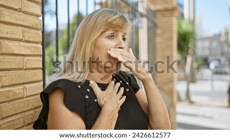 Caucasian blonde, middle age woman looking serious yet relaxed, standing outdoors in sunny city street coughing, showing signs of flu, possibly covid-19, indicating urban infection concern Royalty-Free Stock Photo #2426612577