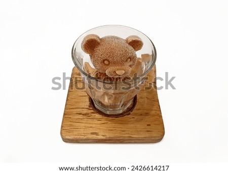 Bear shaped ice cube mold in a cup on a wooden coaster, isolated over white background with cut out. Frozen coffee in the shape of teddy bear. Chocolate milk ice cube in bear shape. Cafe dessert menu.