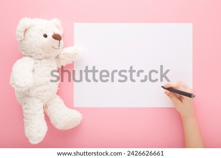 Baby girl hand holding marker and writing greeting card or drawing draw on white paper. White soft teddy bear toy on light pink table background. Pastel color. Closeup. Empty place for text. Top view.