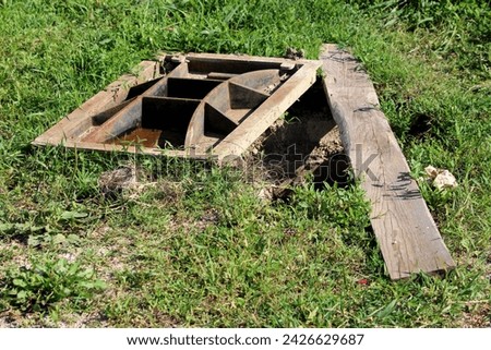 Large heavy custom made rectangle cast metal partially rusted manhole cover next to open manhole temporarily covered with dilapidated wooden board surrounded with uncut grass at local field on warm Royalty-Free Stock Photo #2426629687