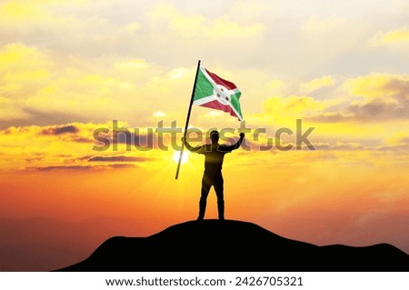 Burundi flag being waved by a man celebrating success at the top of a mountain against sunset or sunrise. Burundi flag for Independence Day.