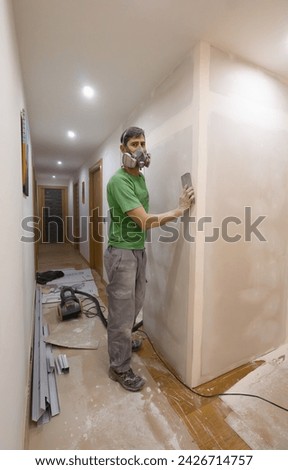 photograph of handyman sanding drywall wall using hand sandpaper and powdered dust mask.