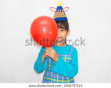 A girl in a birthday hat covers her face partly with a balloon, smiling joyfully.