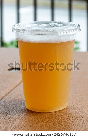 A tall clear plastic glass of sour craft beer in a microbrewery. The cold refreshment has frost on the glass. The beer is on a wooden patio table. The top of the glass has white froth around the rim. 
