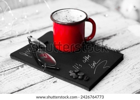 A black and white picture of a hot cup of coffee, on a table with a notebook and glasses.