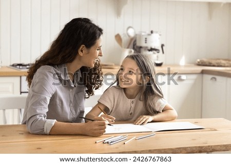 Hispanic loving mom or babysitter spend time with little kid daughter sit at table in kitchen holding colored pencils drawing in paper sketchbook enjoy creative hobby, family activity at home. Leisure