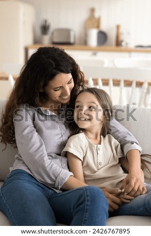 Vertical shot Hispanic woman sit on sofa with pretty daughter, small child enjoy pleasant time with affectionate mommy rest together on couch in cozy flat, smile, lead conversation on weekend at home Royalty-Free Stock Photo #2426767895