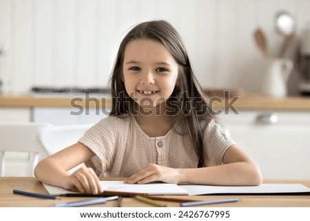 Cute little girl sit at table with sketchbook and colored pencils in cozy kitchen smile staring at camera posing for picture indoors. Children creative hobby, vocation and development, pastime at home