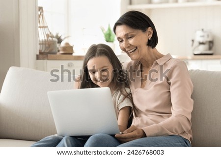 Happy mature grandmother and little cute granddaughter spend time together on internet sit on couch with laptop, watch movies, enjoy funny videos, talking on videocall, having carefree leisure at home