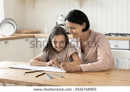 Elderly 60s retired granny spend time with 5s granddaughter sit at table drawing pictures with colored pencils looks interested in creative activity. Children education and skills development, daycare