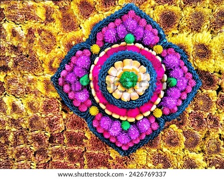 In the background is an orange carnation. In the center of the picture is a flower shape divided by green rope, decorated with pink, purple, yellow, and green amaranths and orange corn seeds.