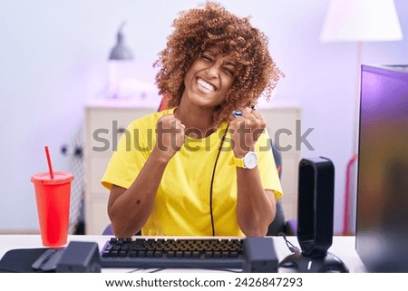 Young hispanic woman with curly hair playing video games wearing headphones very happy and excited doing winner gesture with arms raised, smiling and screaming for success. celebration concept. 