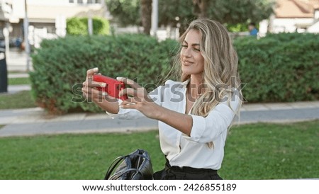Attractive, confident blonde woman joyfully making a fun, casual selfie with her smartphone on a park bench, bathed in beautiful sunlight, her smile radiating pure happiness and positivity.