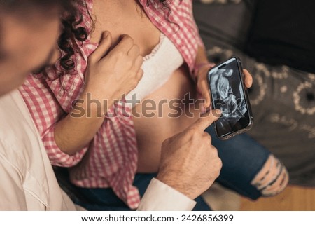 Faceless Pregnant Couple on Sofa Watching And Pointing a Phone with Ultrasound Image. Unrecognizable pregnant couple on sofa, woman showing ultrasound image on phone.