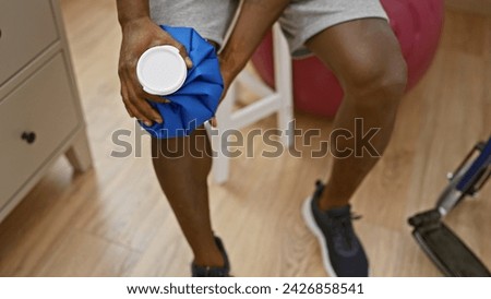 African american man applying ice pack to knee in a rehab clinic room, portraying recovery and healthcare. Royalty-Free Stock Photo #2426858541