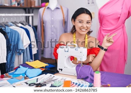 Hispanic young woman dressmaker designer using sewing machine with a big smile on face, pointing with hand finger to the side looking at the camera. 