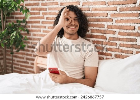Hispanic man with curly hair using smartphone sitting on the bed smiling happy doing ok sign with hand on eye looking through fingers 
