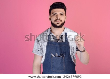 Front view of young hairdresser, designer, barber wearing uniform, standing, showing business card. Isolated on pink studio background.