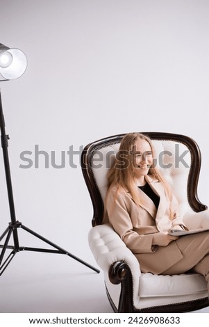 Female, model, blogger, blonde sitting on chair in white photo studio, isolate. Dressed in a formal beige trouser suit