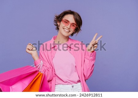Young happy woman with short hair in pink clothes holding colourful shopping bags isolated on violet background. Store, shopping, sales, cyber Monday concept 