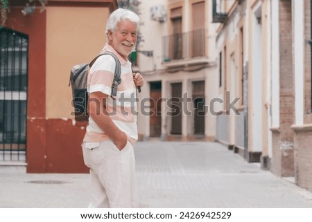 Happy senior tourist man holding backpack on shoulder walking in the alleys of the old town in Seville, Spain looking at camera. Travel and tourism concept Royalty-Free Stock Photo #2426942529