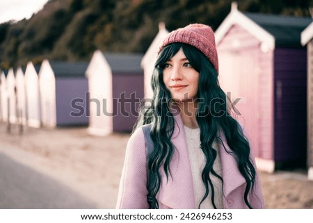 Stylish hipster woman with color hair in pink outfit and backpack walking along wooden beach huts on seaside. Off season Travel concept. Seasonal street fashion. Barbiecore style. Simple pleasures Royalty-Free Stock Photo #2426946253