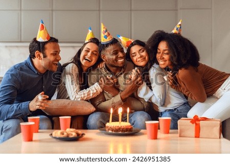 Diverse friends embracing birthday guy sitting with party hats, sharing joy and laughter in heartwarming celebration indoors. Group of multiracial students enjoying bday gathering together