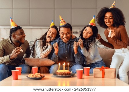 Happy middle eastern guy making wish on his birthday celebration surrounded by diverse friends indoor. Multiethnic students in party hats celebrating bday of their pal in warm and joyful atmosphere