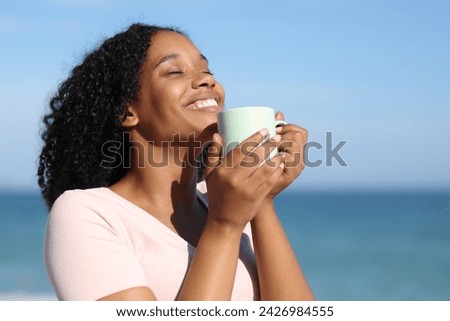 Happy black woman smelling coffee laughing on the beach and smiling Royalty-Free Stock Photo #2426984555