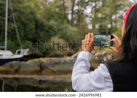 Unrecognizable woman taking photo with mobile phone of a beautiful coastal area with small sailboat protected between a large voice in a sea cove
