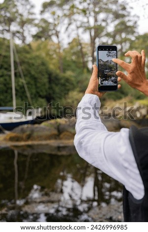 Unrecognizable woman taking photo with mobile phone of a beautiful coastal area with small sailboat protected between a large voice in a sea coveBe