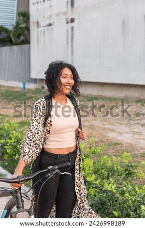 Latin woman with curly hair stands with her bicycle, sporting a leopard print jacket and backpack.