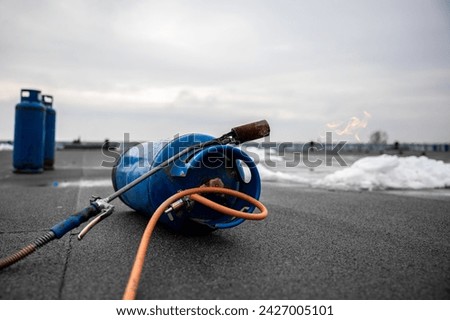 gas torch tool lying on new roofing surface with propane tank and hose, essential equipment for construction workers for melting and sealing tar on flat roof Royalty-Free Stock Photo #2427005101