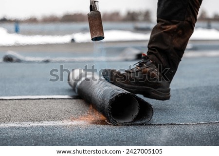 professional roofer at work securing rolled tar paper with a blowtorch on a flat roof installation process Royalty-Free Stock Photo #2427005105