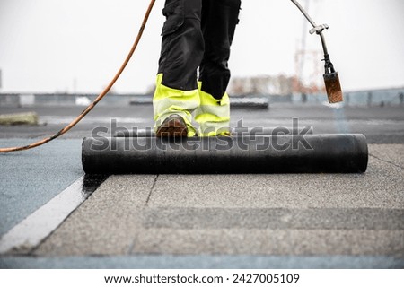 professional roofer applying tar with blowtorch on new roofing felt for waterproofing a flat roof construction site with safety gear Royalty-Free Stock Photo #2427005109