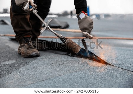 professional roofer applying bitumen roll on flat roof with a gas burner at a modern construction site Royalty-Free Stock Photo #2427005115