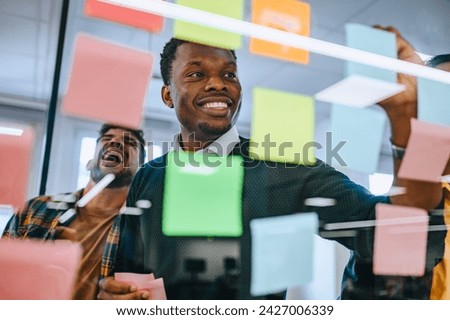 Portrait of a smiling african american businessman working on a new project and writing main topics while using colorful sticky notes on the glass wall. Shot trough a glass wall. Copy space.
