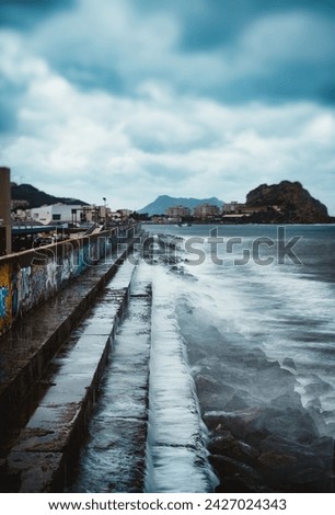 "Coastal area, cloudy sky, waves, graffiti wall, buildings in background."