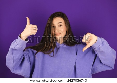 Young woman in purple hoody making good-bad sign isolated on violet background. Girl make choice, or makes a decision thumb up or thumb down, like or dislike, yes or no. Royalty-Free Stock Photo #2427036743