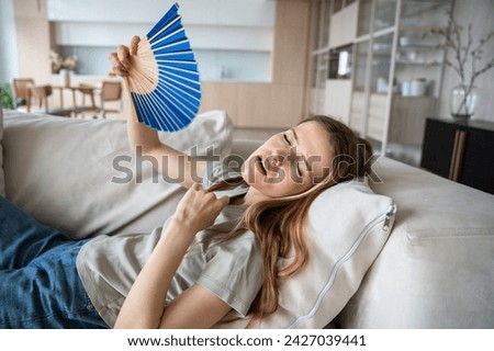 Exhausted weary woman lying on sofa with waving paper fan, suffering from high temperature in apartment with broken conditioner. Overheated sweating young female feeling unwell, dehydrated trying cool Royalty-Free Stock Photo #2427039441