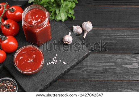 Flat lay composition with delicious ketchup, spices and products on black wooden table, space for text. Tomato sauce