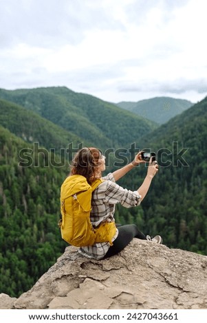 Tourist woman taking picture outdoors for memories, making selfie on top of cliff with valley mountains view, sharing travel adventure journey. 