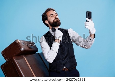 In studio, bellman in uniform takes pictures while using mobile device for selfies and acting foolish against blue background. Classy hotel porter does photos with his phone while being on duty.