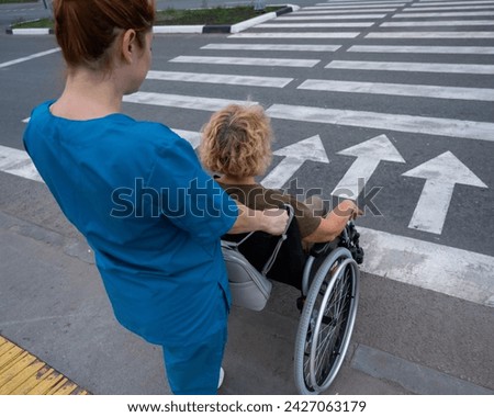 Rear view of a nurse helping an elderly woman in a wheelchair cross the road.  Royalty-Free Stock Photo #2427063179