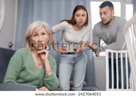 Sad mature woman sitting at home while emotional young adult couple berating her, family relationship problems concept