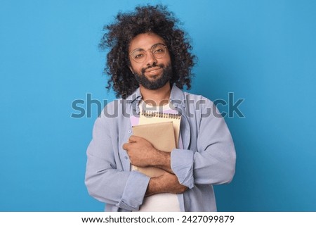 Young optimistic smart Arabian man with glasses holds several notebooks to prepare for exam or test that allows you to become member of university community of talented students stands in blue studio Royalty-Free Stock Photo #2427099879