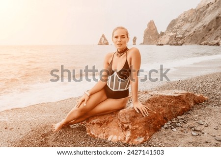 Woman travel portrait. Happy woman with long hair poses on a red volcanic rock at the beach. Close up portrait cute woman in black bikini, smiles at the camera, with the sea in the background.