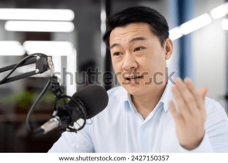 Close-up photo of an Asian young man sitting in the office at a desk in front of a microphone and having an online meeting via video call, discussing, explaining, gesturing with his hand.