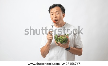 Portrait of an Indonesian Asian man, wearing a white T-shirt, joyfully posing while eating salad from a large transparent bowl, isolated against a white background.